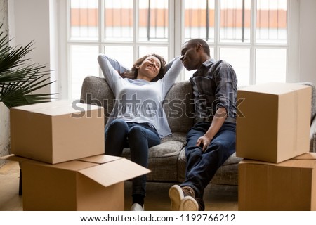 Black married couple sitting on couch in living room at house. Smiling happy wife and husband relaxing resting unopened belongings still in their cardboard boxes. Moving and relocate new home concept