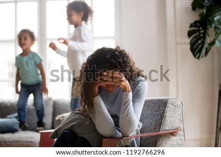 Black single frustrated woman hold her head with hands sitting on chair in living room, playful kids jumping on couch on a background. Tiredness, depression difficult to educate children alone concept