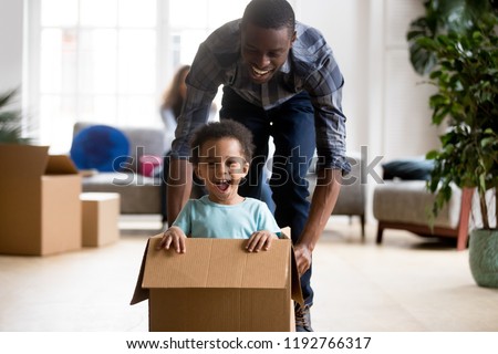 Black family in living room have a fun spend time at new home. African adorable playful laughing boy sitting at cardboard box, father rolling him playing together. New property and relocation concept