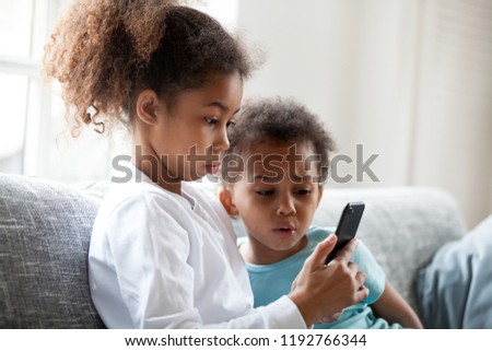 African brother and sister sitting on couch at home using mobile phone.Close up surprised daughter and frustrated son looking at device screen. Technology new generation addicted with gadgets concept