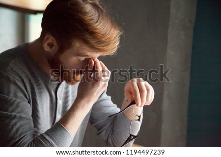 Tired red haired man take glasses off working too long at computer, exhausted millennial male suffer from headache or eye tension, young student massaging nose bridge feeling fatigue sitting in café