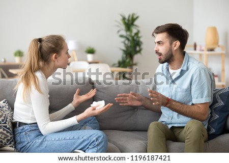 Unhappy young millennial married couple sitting on couch in living room at home and quarrelling. Husband and wife emotionally arguing talking cheating. Break up and problems in relationships concept