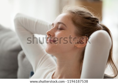 Close up portrait serene woman smiling sitting on couch at home. Girl has a break after work or study closing eyes putting hands behind head relaxing thinking, feels happy breathing fresh air concept