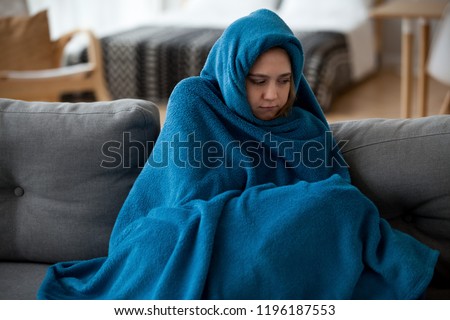 Young unsatisfied sick woman covered with warm blue blanket plaid from head to toes sitting alone on couch in living room at cold home. Seasonal problems, virus diseases and central heating concept