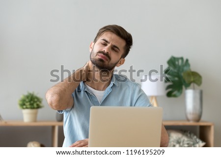 Millennial tired stressed man having a neck pain. Male touching massaging neck sitting at the desk working or studying indoors, suffering from discomfort long hours of sedentary overworking concept