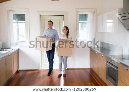 Full length millennial wife and husband hold carton boxes with belongings standing in modern kitchen at home. Attractive happy couple bought new house. Moving relocate day mortgage and tenant concept