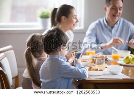 While whole family eating talking having breakfast, little preschool son in glasses holding using smartphone looking at screen. Bad habit overuse of devices and gadgets, mobile phone addiction concept