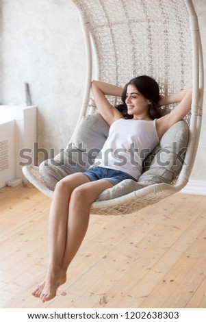 Smiling young barefoot woman sitting, relaxing with hands behind head in lounge hanging chair, looking in distance, dreaming about good future, happy girl resting in cozy, comfortable sofa at home