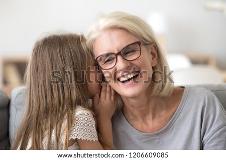 Cute small granddaughter whisper something in granny ear, grandmother and grandchild share secrets having sincere talk at home, grandma and little girl have fun at home together laughing
