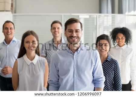 Smiling leader ceo or professional business coach looking at camera posing in office with diverse happy team at background, successful startup founder, corporate employee with staff members portrait