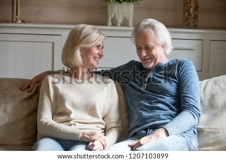 Happy aged couple sit on cozy couch at home talking or having conversation, senior husband and wife enjoy leisure time relaxing on sofa together, smiling elderly man and woman hug and cuddle