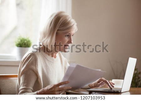 Senior woman holding papers busy at laptop managing house utility bills or finances, aged female using computer working with bank loan or mortgage documents online. Elderly and technology concept