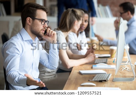 Angry male employee confused working at computer, solve business problems on phone, mad worker talk on cell arguing with client or customer, man busy at pc having serious conversation on cellphone