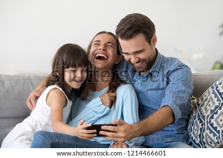Smiling happy multi-ethnic family sitting on couch in living room at home. Mother father and little daughter laughing using mobile phone watching funny videos online have fun looking humorous program