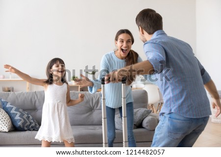 Happy couple and small daughter standing in living room at home. Cheerful wife little girl meets husband father he arrived at home with suitcase luggage from long business trip. Family reunion concept