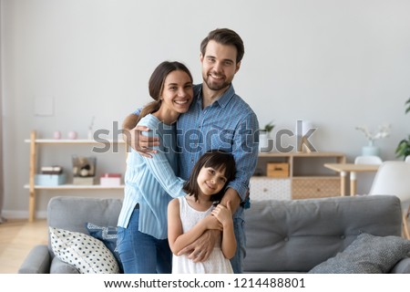 Cheerful diverse multi-ethnic family married couple wife husband little daughter embracing standing together in living room smiling looking at camera at new modern home feels happy and satisfied