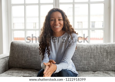 Head shot portrait attractive African American woman, making video call, using computer, sitting on couch, sofa in living room, happy young female smiling, look at camera