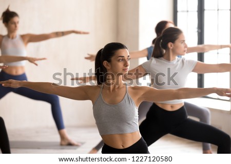 Indian girl and diverse group of young people practicing yoga lesson doing Warrior II exercise, Virabhadrasana 2 pose, working out, indoor close up, female students training at sport club or studio