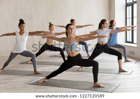 Indian girl and diverse group of young sporty people practicing yoga, doing Warrior II exercise, Virabhadrasana 2 pose, working out, indoor full length, female students training at sport club, studio