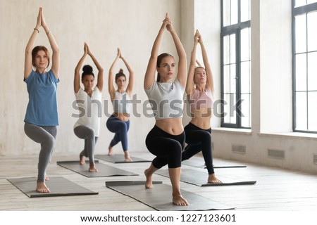 Group of young sporty people practicing yoga lesson, doing Warrior one pose, Virabhadrasana 1 exercise, working out, indoor full length, mixed race students training at sport club or yoga studio