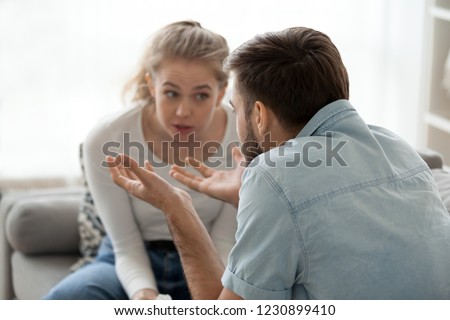 Millennial couple arguing sitting on couch at home, emotional husband gesturing proving his point of view to stubborn wife, man and woman dispute having fight or disagreement. Family conflict concept