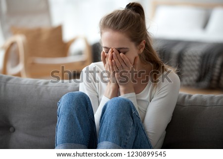 Upset young female sit on couch at home crying over ended relationships, unhappy girl cover face feeling down after breaking up with boyfriend, woman lie on sofa have problems in life