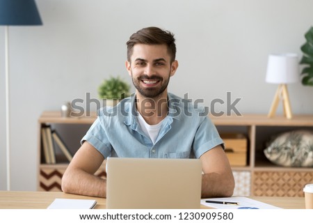 Portrait of happy millennial man sitting at office desk working with laptop and paperwork from home, headshot of smiling male student posing at workplace using computer studying looking at camera