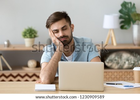 Exhausted millennial male hold head with hand sitting at office table falling asleep, tired man fall asleep working at laptop at home workplace, sleepy guy feel fatigue taking nap near computer