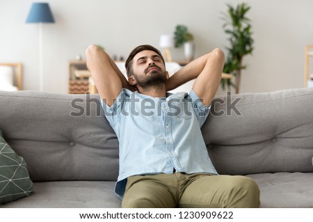Calm millennial man relax on cozy couch hands over head sleeping or taking nap, peaceful male lying on sofa with eyes closed having rest or visualizing, tired guy fall asleep chilling at home