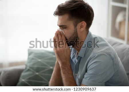 Ill millennial man sit on couch feel unwell blowing running nose with napkin, sick male at home have health problems, get flu or fever symptoms, tired guy suffer from sickness sneezing holding tissue