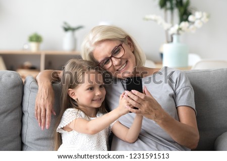 Happy grandmother and cute granddaughter using cellphone together, smiling older grandma and child girl having fun taking selfie on phone, cheerful granny with little kid play making photo on mobile