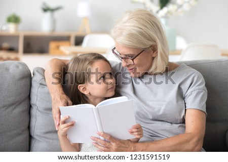 Loving grandmother teaching granddaughter holding book sitting on sofa, grandma baby sitter embracing kid girl reading fairytale to cute child, nanny granny telling story to preschool grandchild