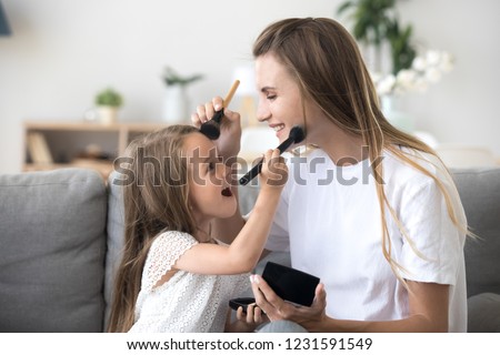 Smiling mom and kid preschool daughter doing makeup together, excited little girl holding make-up brush puts powder on mothers face, happy child applying blush having fun playing with mommy at home