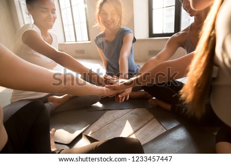 Young diverse girls start new day with sport gathered at fitness studio sitting in circle on yoga mats putting their hands together showing unity teamwork and support, sunlight through the big windows