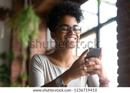 Smiling happy African American woman using phone, browsing mobile apps or websites, get good message from friend or boyfriend, check social network, looking at smartphone screen, reading good news