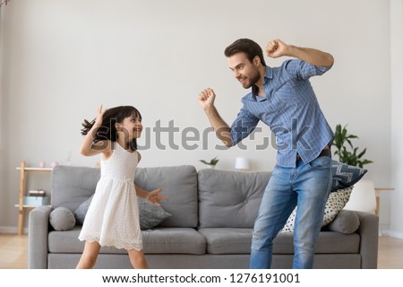 Happy dad and little funny daughter laughing dancing in living room, cheerful daddy and cute child girl having fun together, active kid enjoying copying fathers moves playing with parent at home