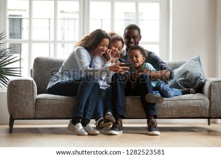 Happy african family with 2 children son daughter having fun with gadget on couch using smartphone at home, black parents and kids laugh watch funny video, make video call selfie on phone online app