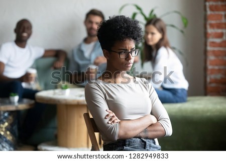 Unkind teenagers looking laughing at black classmate, focus on mixed race unhappy shy and frustrated girl wearing glasses sitting at closed posture arms crossed feeling insecure having low self-esteem