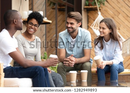 Four laughing diverse people positive multi-ethnic friends sitting in cozy cafe summer terrace drink coffee telling funny stories from life feels happy and satisfied enjoy time together on weekend