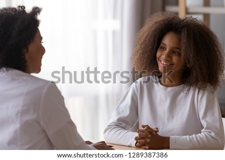 Smiling African American teenage girl sit at table talking with female teacher or tutor at home, happy teenager have casual conversation with woman doctor or specialist. Adult children relationships