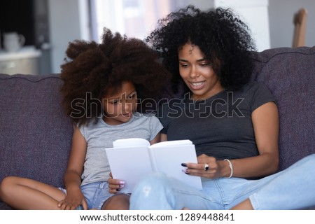 African mom holding book telling story to little daughter spending time at home, child girl enjoying listening to fairy tale sitting on couch with black mother, kid learning reading, family activity