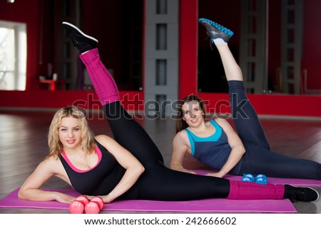 Two smiling pretty girls do leg swings, exercises for hips and buttocks on mats in sports hall