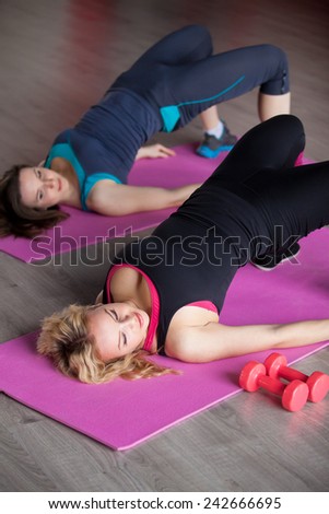 Two pretty girls do fitness exercises for hips and buttocks in lying position on mats in sports hall