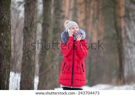 Happy beautiful girl in knitted hat and red winter coat, outdoors, in park, using mobile phone, talking on smart phone, answers the call