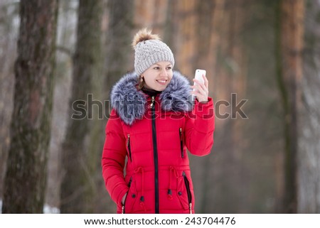 Happy beautiful girl in knitted hat and red winter coat, outdoors, using mobile phone, looks on the screen of smart phone