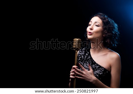 Beautiful young female vocalist in shiny black evening dress singing with emotions behind golden retro microphone, during live musical show, copy space