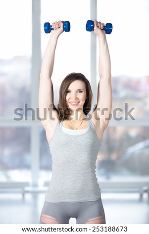 Sporty smiling beautiful female athlete doing fitness exercises in well-lit fitness center with dumbbells, weight training, warming up