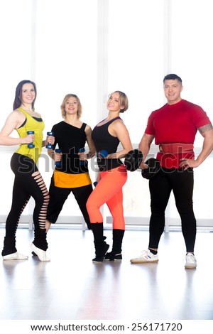 Team of positive sporty people, friends, coaching staff in fitness center holding dumbbells. Muscular powerlifter and three young female athletes lifting weights in gym