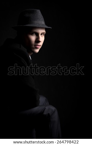 Low key shot of mysterious young man wearing hat and coat sitting in the darkness, copy space