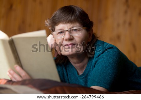 Beautiful elder woman in spectacles reading a book with faint smile, lying relaxed on bed at home in comfy bedroom with wooden walls, absorbed in interesting novel storyline